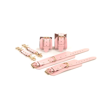 Pink leather BDSM set laid flat: wrist cuffs and ankle cuffs with buckle straps, rivets, adjustment holes, and gold-tone hardware. A 2.5 cm D-ring is visible on each cuff. Two leather connectors with gold-tone carabiners on both ends are placed beside the set.
