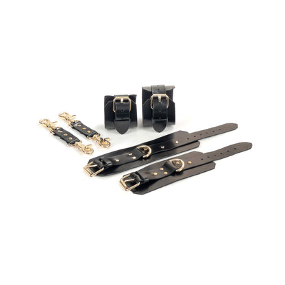 Black leather BDSM set laid flat: wrist cuffs and ankle cuffs with buckle straps, rivets, adjustment holes, and gold-tone hardware. Each cuff shows a 2.5 cm D-ring for attachment. Two matching leather connectors with gold-tone carabiners on both ends are placed to the left.
