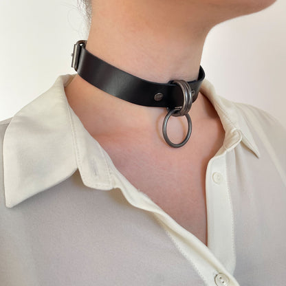 Black leather choker worn in a semi-profile view with dark nickel D-rings and O-ring, matte black strap sitting close to the neck with a straight cut edge, darker metal hardware creating a subdued, graphic look.