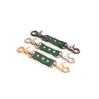 Leather double carabiner connector made of green genuine leather with metal carabiners fixed on both ends, shown on a white background. The image displays green leather combined with gold, silver, and dark nickel carabiners, with the strap laid flat and both carabiners fully visible from above.