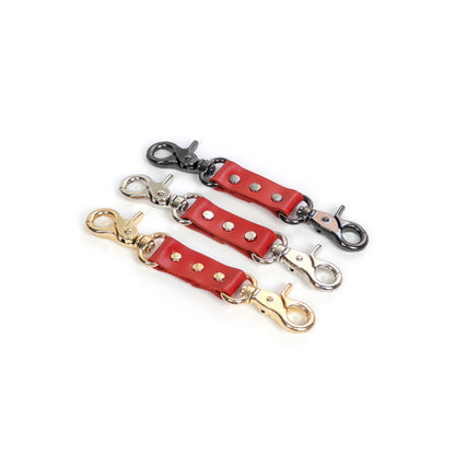 Leather double carabiner connector made of red genuine leather with metal carabiners fixed on both ends, photographed on a white background. The image shows red leather combined with gold, silver, and dark nickel carabiners, with the connector positioned horizontally and all attachment elements clearly visible.