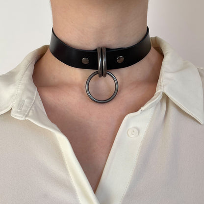 Front view of a black leather choker with dark nickel hardware, two D-rings holding a round O-ring at the center, leather strap evenly aligned around the neck with a straight cut edge, minimal balanced design