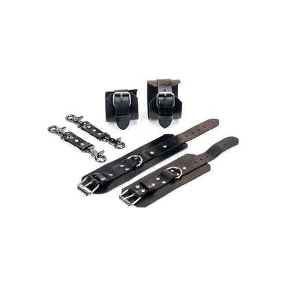Black leather BDSM set laid flat: wrist cuffs and ankle cuffs with buckle straps, rivets, adjustment holes, and dark nickel hardware. A 2.5 cm D-ring is visible on each cuff. Two matching leather connectors with dark nickel carabiners on both ends are placed to the side.