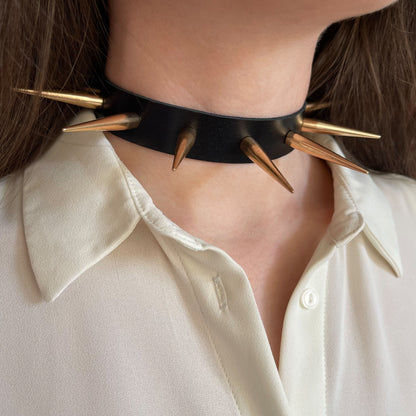 Minimalist black leather choker with large, glossy gold-tone metal spikes. Side view highlights the aggressive yet elegant design, worn over a white blouse. The spikes form a symmetrical halo around the neck, creating a strong, fashion-forward look.