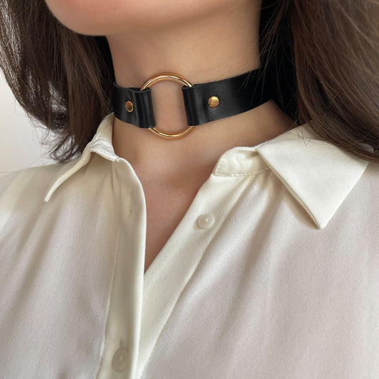 Black leather choker worn on the neck, shown from a semi-side angle. The strap is 20 mm (0.79 in) wide, made of smooth natural leather with a semi-gloss finish and moderate firmness. A large round gold-tone metal ring, 30 mm (1.18 in) in diameter, is placed at the center and fixed with gold-tone rivets. The choker follows the neck line evenly.