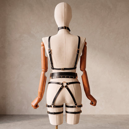 A cream-colored fabric mannequin with wooden arms is shown from the back, wearing a black leather harness-style bodysuit with gold-tone hardware against a textured beige studio background.