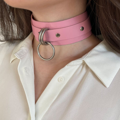 Wide pink leather choker with silver hardware worn in a semi-profile view. The 40 mm wide leather band sits firmly on the neck, with a round O-ring suspended between two D-rings at the front. The narrower 20 mm strap continues toward the back.