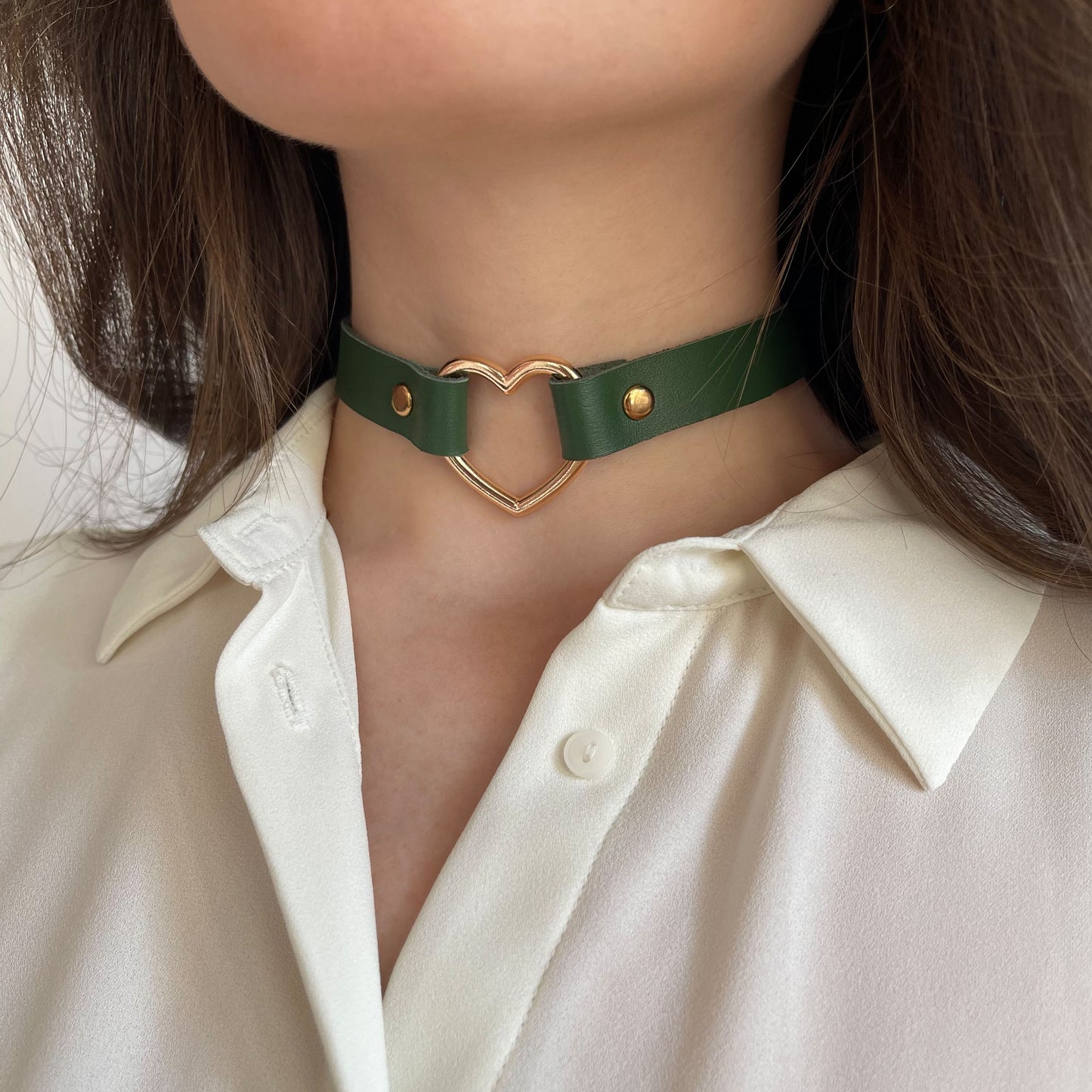 Green leather choker worn on the neck, shown from a semi-side angle. The 15 mm (0.59 in) wide strap is smooth to the touch and moderately firm. A gold-tone heart-shaped metal ring, 30 mm (1.18 in) in diameter, is fixed at the center with gold rivets. The choker rests evenly against the skin.
