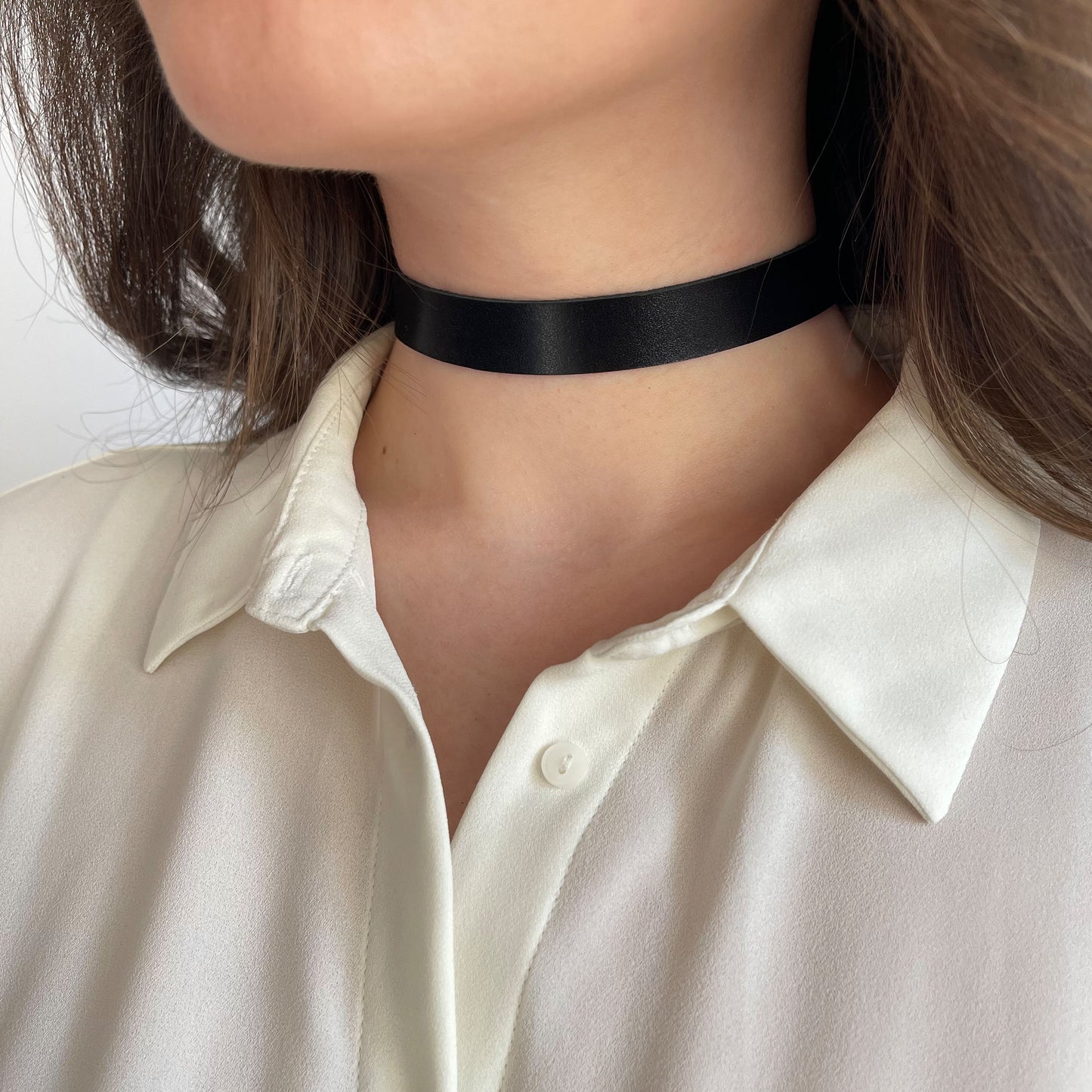Classic black leather choker crafted from semi-matte genuine leather, 1.5 cm (0.59 in) wide and 1.4–1.8 mm thick. Fastens with a sturdy buckle. Worn snugly around a woman’s neck in a light shirt, creating strong visual contrast. Versatile minimalist piece for daily wear or a subtle evening accent.