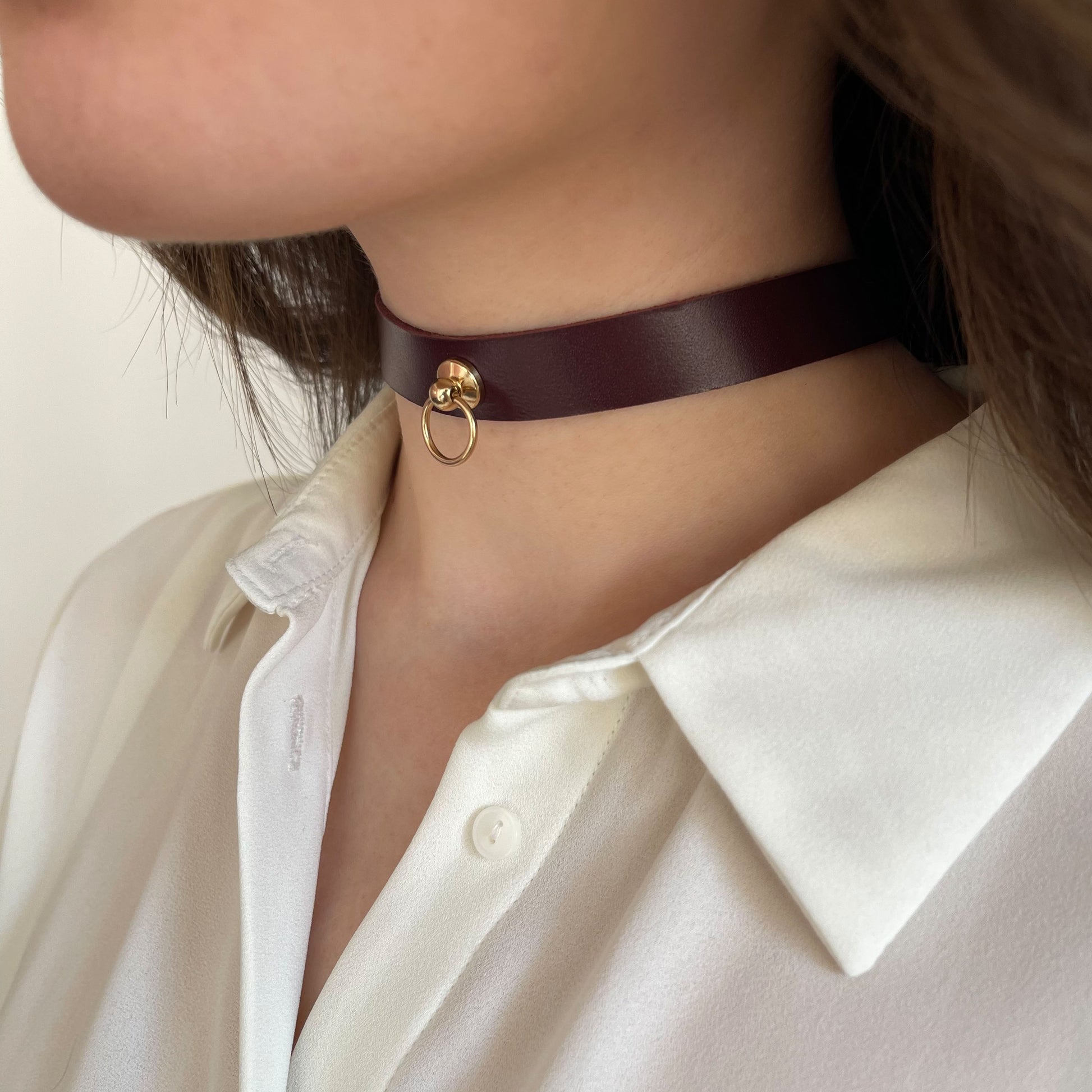 Elegant marsala leather choker with a golden center ring. Made of genuine semi-matte leather (1.4–1.8 mm thick), 1.5 cm (0.59 in) wide. Side view of a woman’s neck in a white blouse, emphasizing the deep color and clean minimalist silhouette of the accessory.