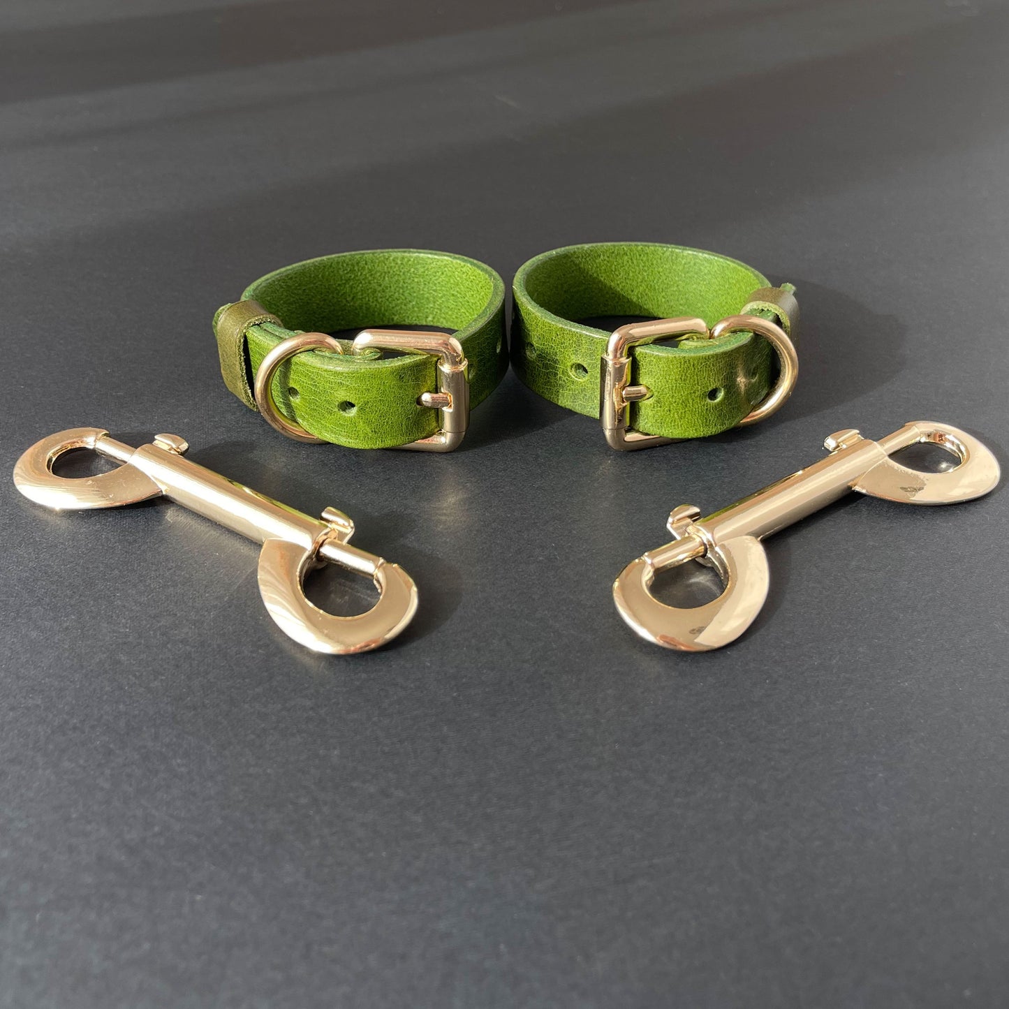 Two green leather handcuffs with brass hardware on a gray background