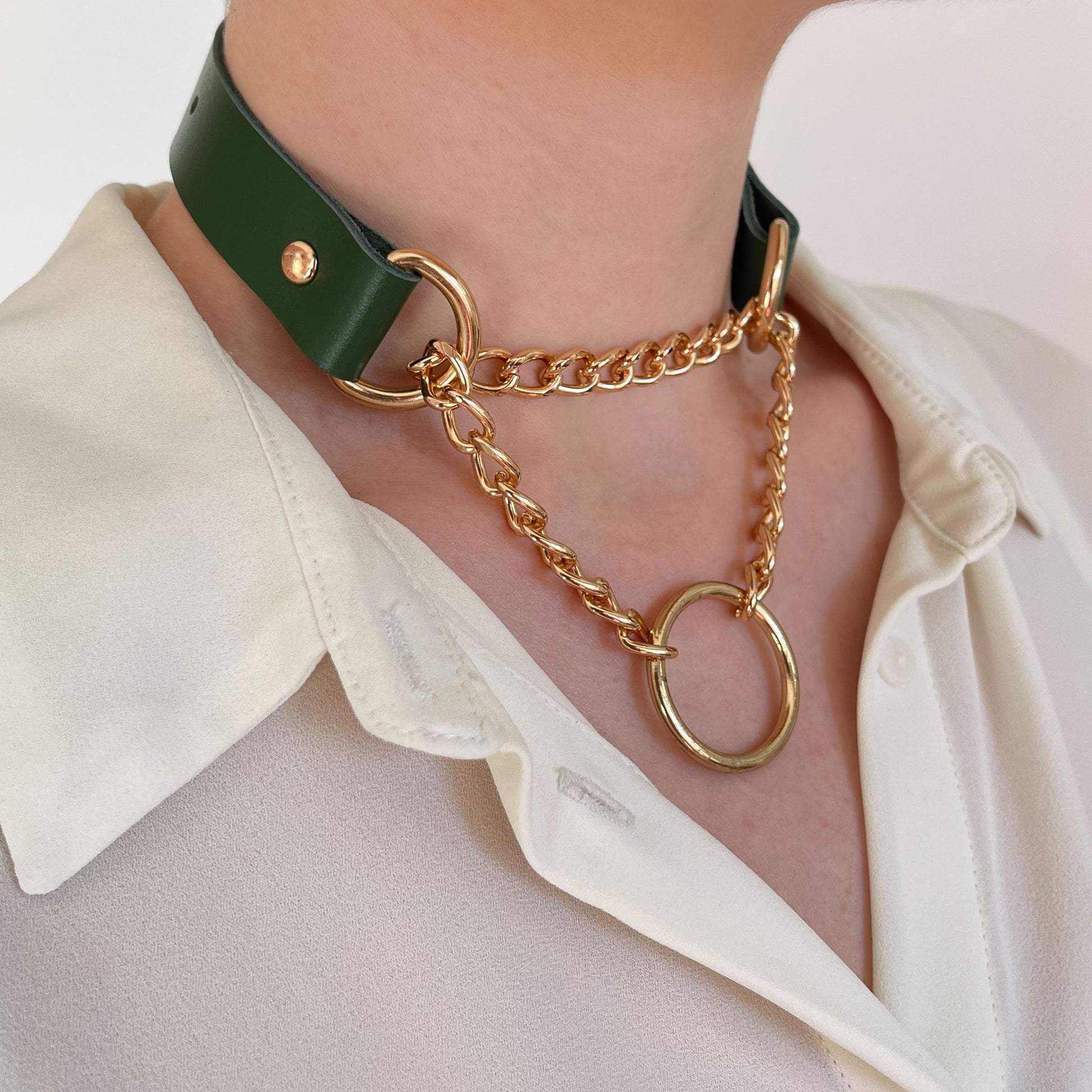 Green leather choker with gold hardware and O-ring, worn over a cream shirt. Decorative layered chains.
