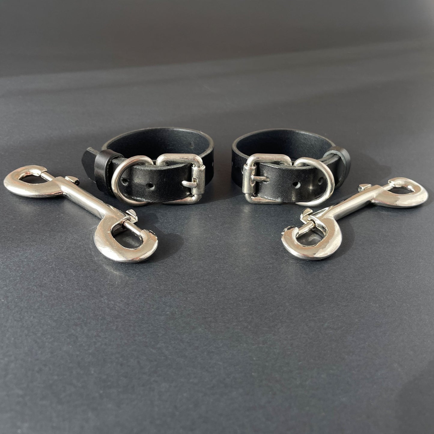 Two black leather slim handcuffs with metal buckles and hooks on a gray background