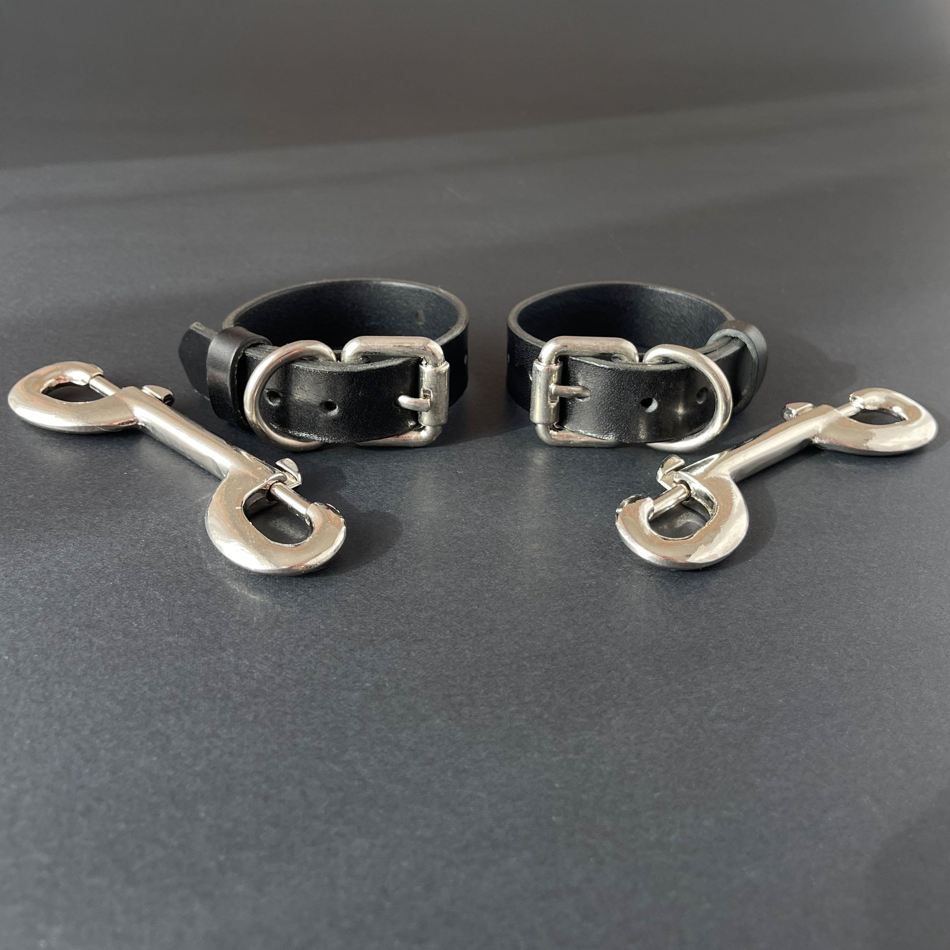 Two black leather slim handcuffs with metal buckles and hooks on a gray background