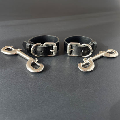 Two black leather slim handcuffs with metal buckles and hooks on a gray background