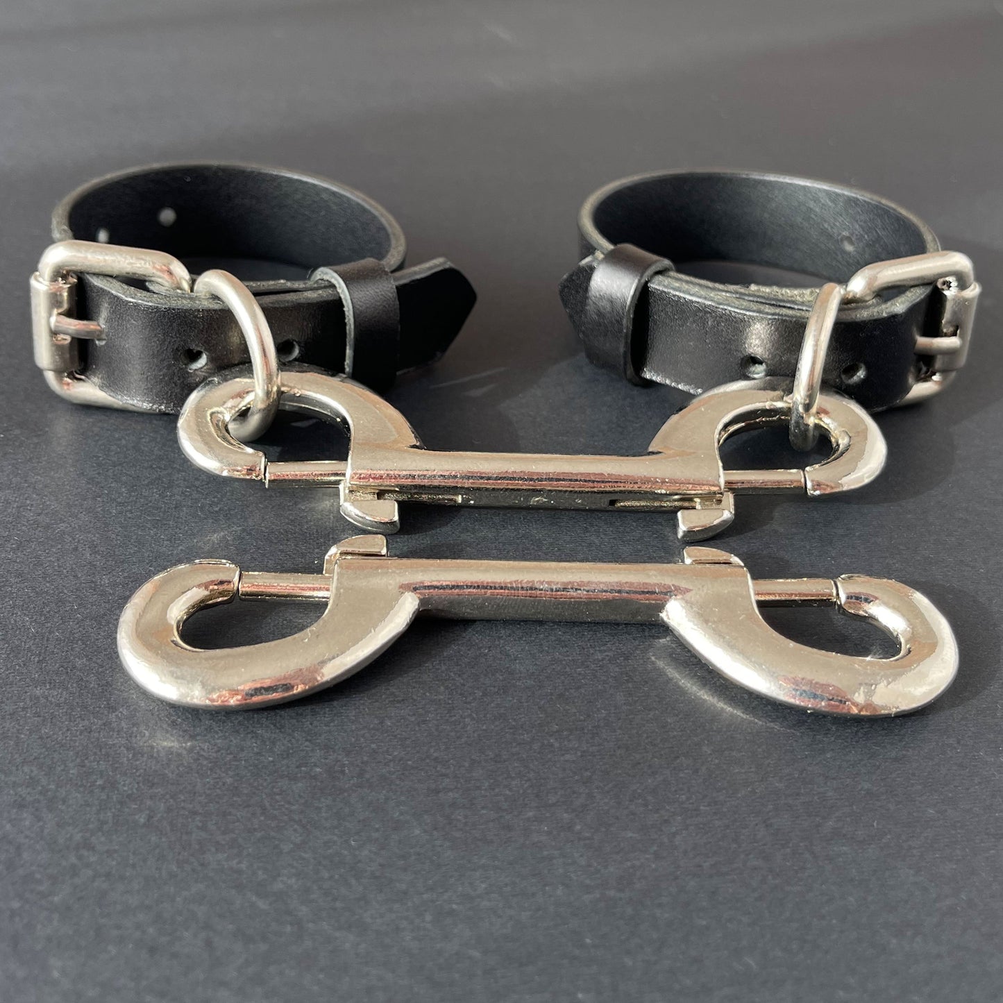 Two black leather handcuffs with silver metal clasps on a dark surface