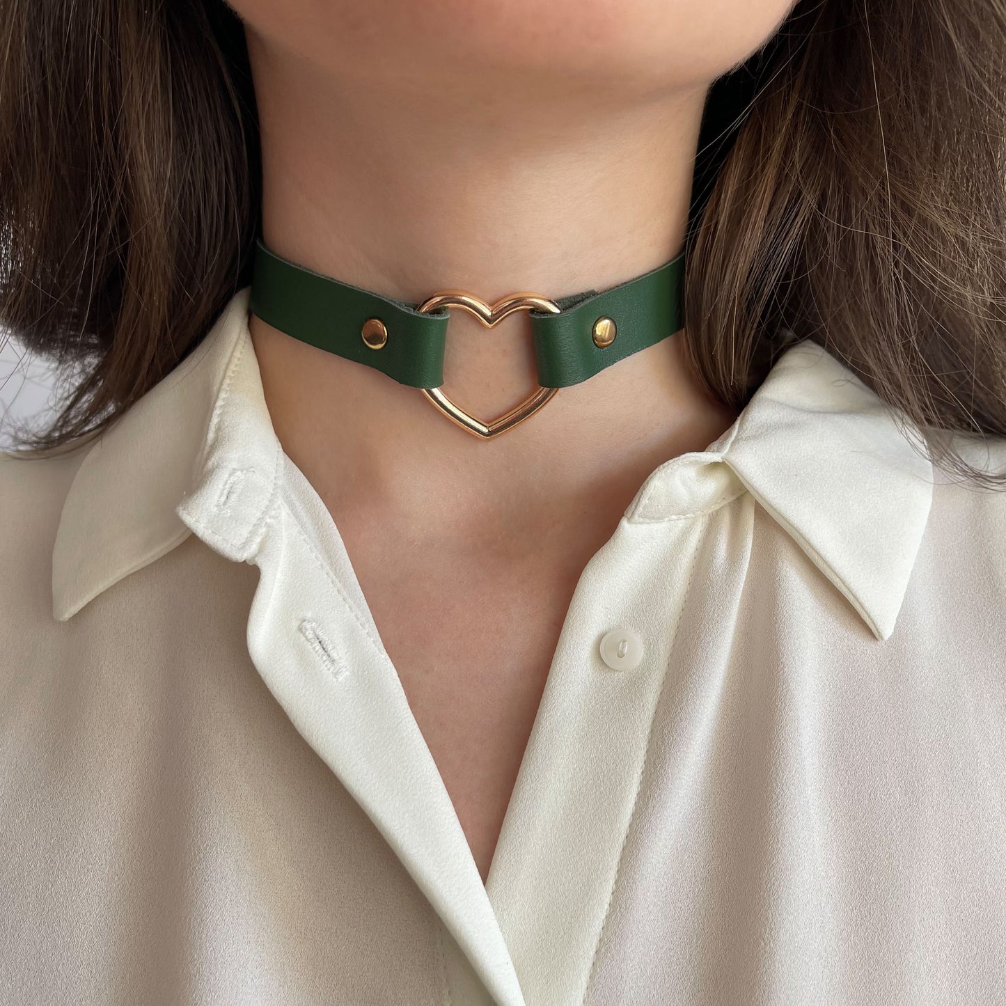 Green leather choker shown from the front while worn close around the neck. The slim leather strap is 15 mm (0.59 in) wide and lies flat. A gold-tone heart-shaped metal ring with a 30 mm (1.18 in) diameter is centered at the front, forming a gentle focal point suitable for everyday wear.