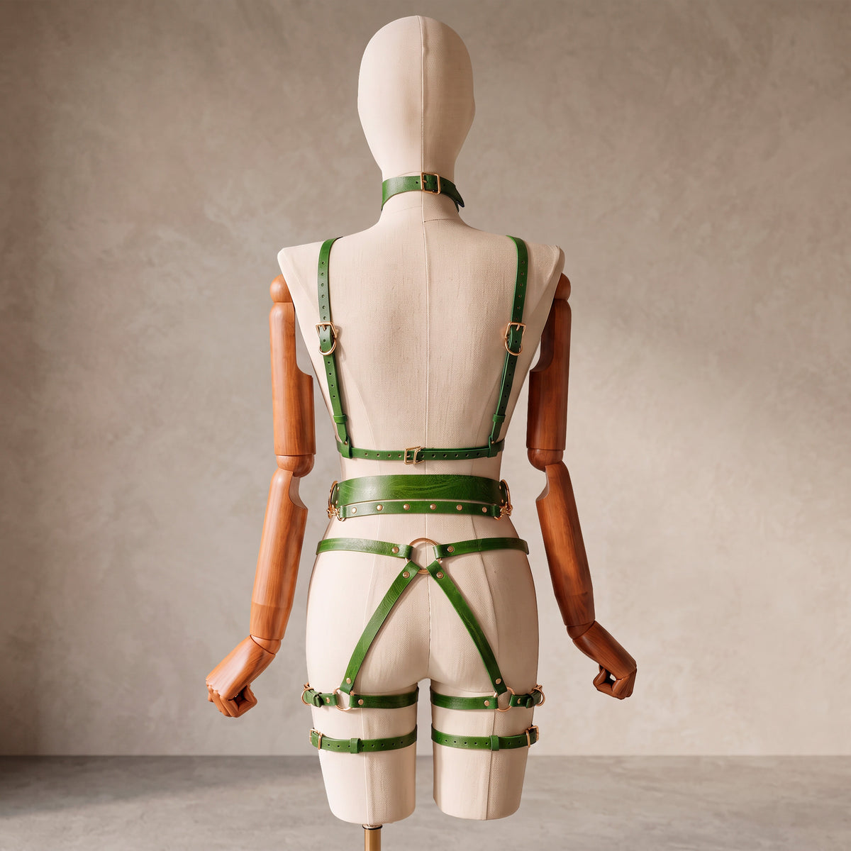 A green leather harness-style outfit with matching belt and choker, displayed on a cream fabric mannequin with wooden arms against a neutral studio backdrop.