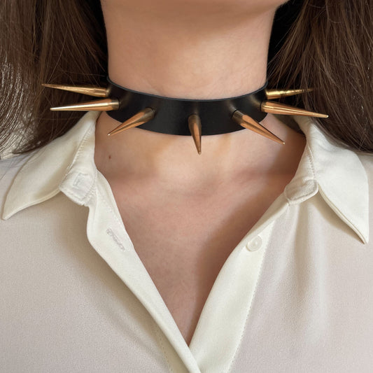 Front view of a wide black leather choker with gold-tone metal spikes. The bold and edgy silhouette stands out against the white fabric. Designed for alternative fashion, this collar delivers dramatic visual impact with every outfit.