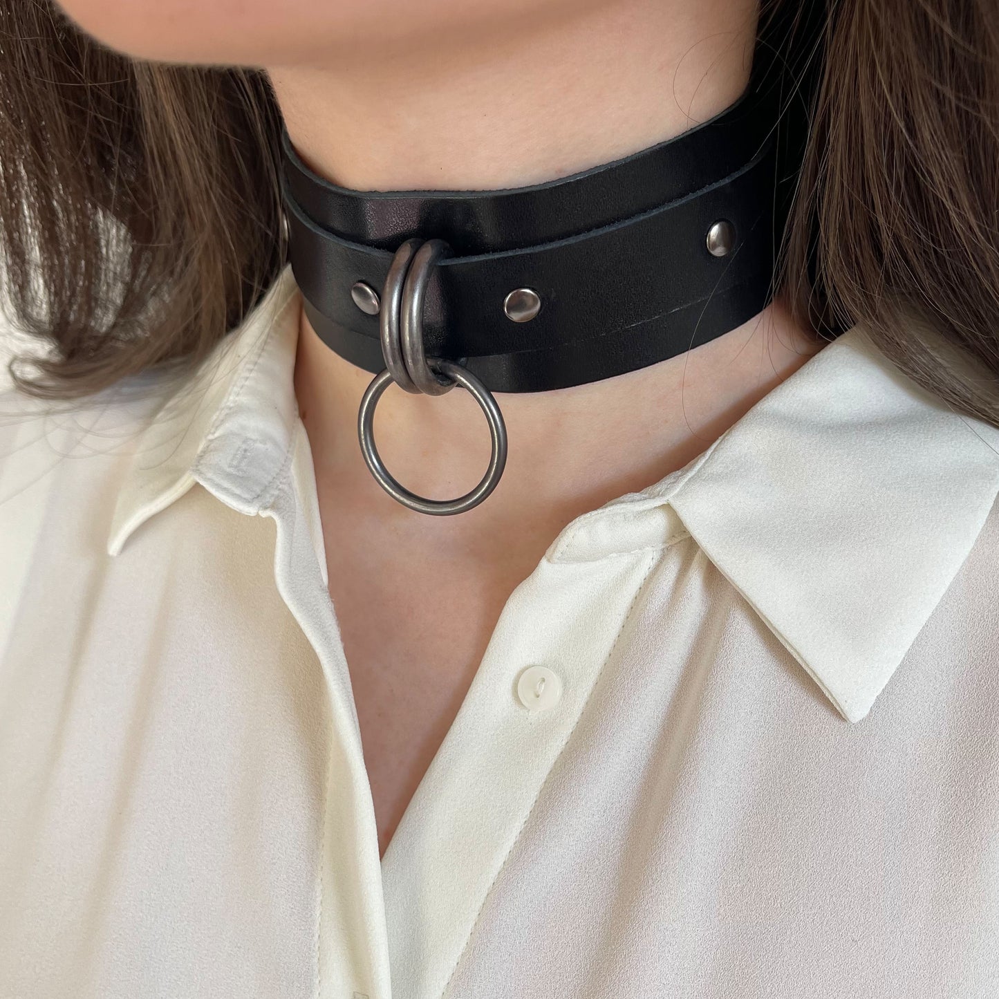 Wide black leather choker with dark nickel hardware shown in a semi-profile view. The 40 mm wide leather band wraps around the neck, with two D-rings and a round O-ring forming the front focal point. The matte metal hardware contrasts with the leather.