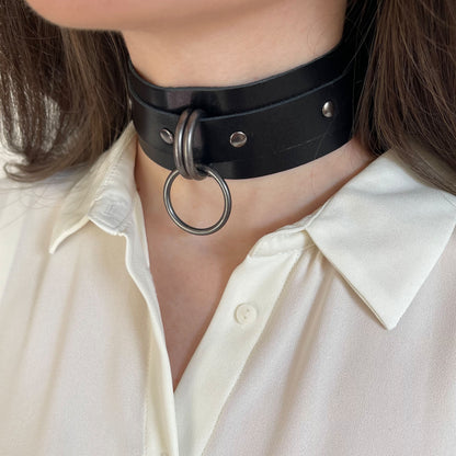 Wide black leather choker with dark nickel hardware shown in a semi-profile view. The 40 mm wide leather band wraps around the neck, with two D-rings and a round O-ring forming the front focal point. The matte metal hardware contrasts with the leather.