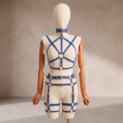 A cream-colored mannequin torso with wooden arms and legs is shown from the back, wearing a blue leather harness-style outfit with multiple straps, buckles, and rings against a textured beige studio background.