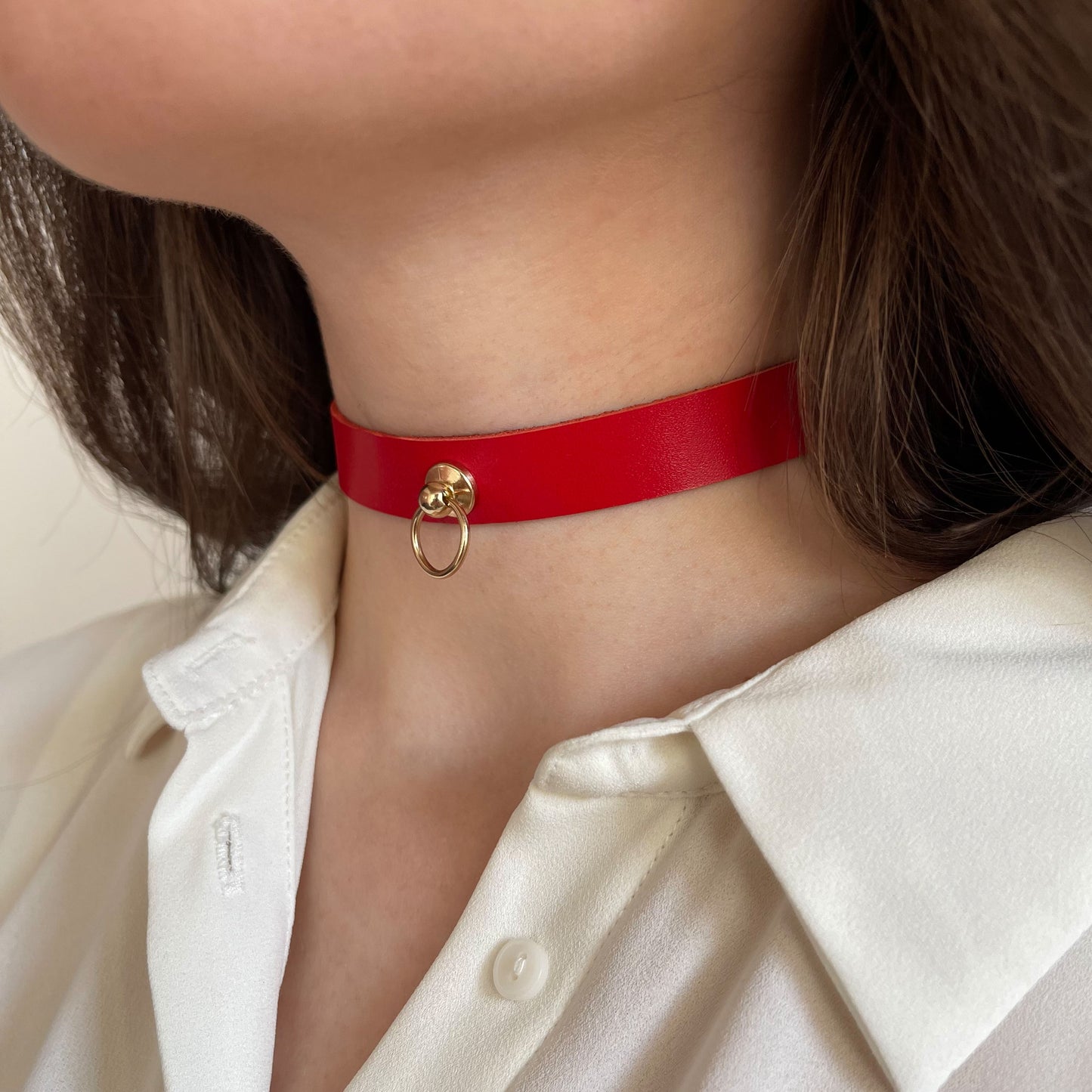 Red leather choker worn on the neck, shown from a semi-side angle. The strap is narrow, smooth, and semi-glossy, made from natural leather. At the center front, a small metal ring is fixed with a screw rivet. The choker rests evenly against the skin.