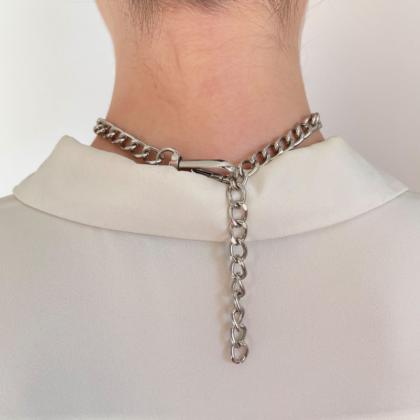 Close-up of a silver-tone choker clasp with a dangling extender chain. Back of the neck view with a soft white collar. Functional and stylish, this fastening detail complements modern metallic necklaces and provides an adjustable fit.