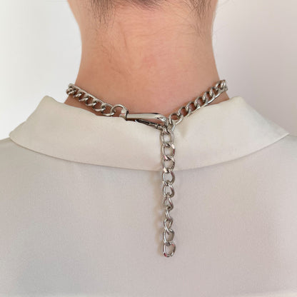 Close-up of a silver-tone choker clasp with a dangling extender chain. Back of the neck view with a soft white collar. Functional and stylish, this fastening detail complements modern metallic necklaces and provides an adjustable fit.