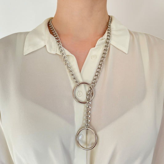 Silver-tone sliding chain choking collar worn on the neck over light clothing. One 30 mm (1.18 in) ring sits closer to the neck, while the second ring hangs lower. The metal chain passes through the upper ring, clearly showing the sliding tightening mechanism.