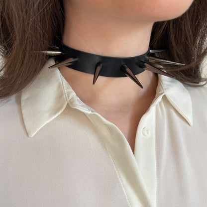 Side view of a black leather choker featuring glossy black cone spikes. Worn on a woman’s neck with a soft white blouse, this accessory captures bold contrast and gothic minimalism in a clean, streamlined form.