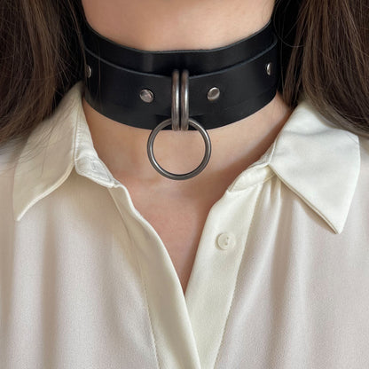 Front view of a wide black leather choker with dark nickel hardware worn on the neck. The round 25 mm O-ring is centered between two D-rings on the wide leather band. The straight cut leather edges and solid metal elements are visible.