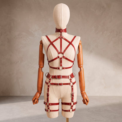 A cream-colored mannequin with wooden arms and hands displays a red leather harness-style bodysuit from the back, featuring multiple straps, rings, and buckles against a textured beige studio backdrop.
