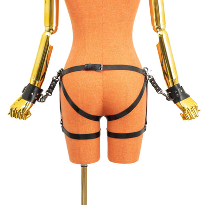 Back view of an open crotch hip ring garter belt set on a mannequin in black leather, showing the back waist buckle, side hip rings, straps running under the buttocks and through the crotch, lower leg rings, thigh straps with buckles, and wrist cuffs attached at both sides.