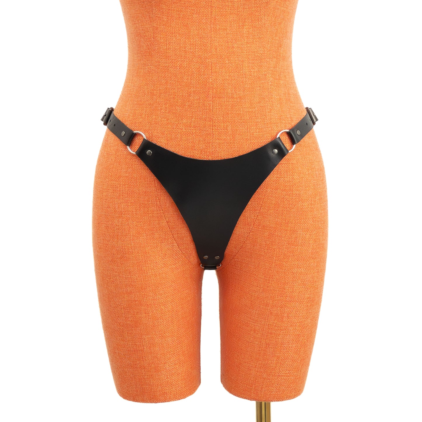 Front view of black leather thong on orange mannequin showing the wide front panel with ring connections and side buckle straps forming a defined structured shape.