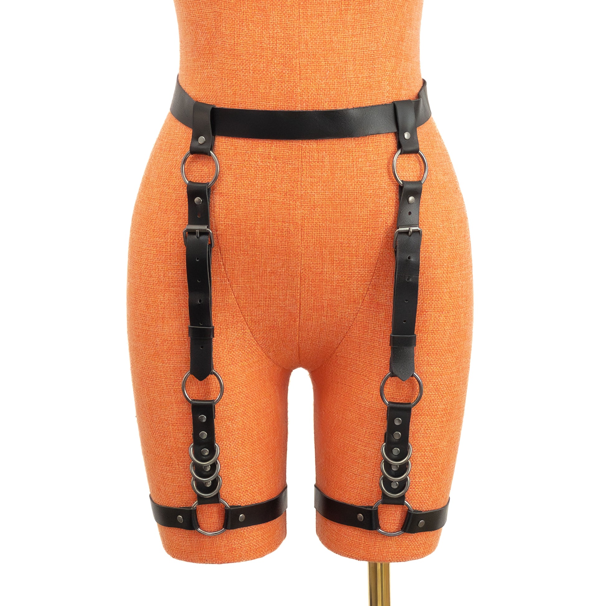 Three-quarter front view on a mannequin showing the garter belt from an angle. The black leather waist strap, vertical straps, and thigh bands are connected with dark nickel O-rings and D-rings. Buckles and strap ends are visible along the outer thigh.