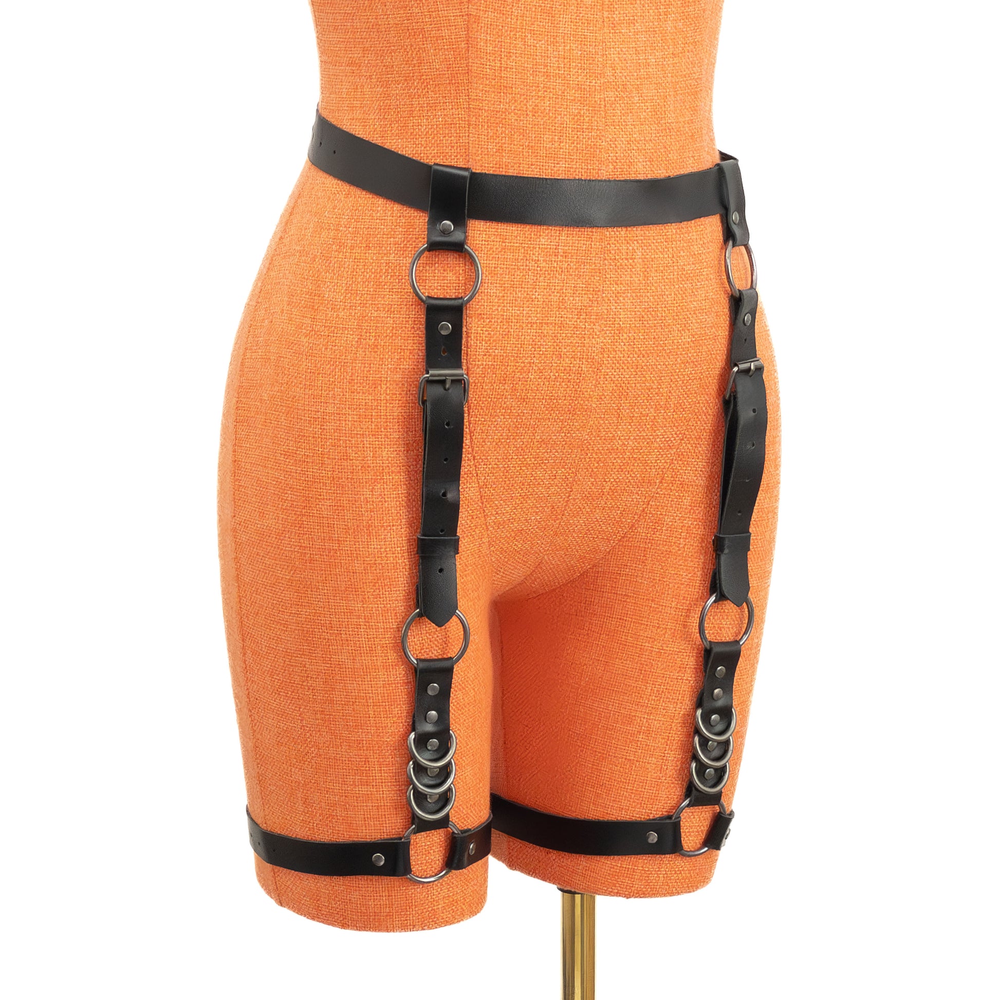 Front view of the garter belt on a mannequin. Black leather waist strap connects to vertical straps ending at thigh bands. Dark nickel O-rings and D-rings are visible on each side, with metal buckles and stacked rings near the thighs showing the full front layout.