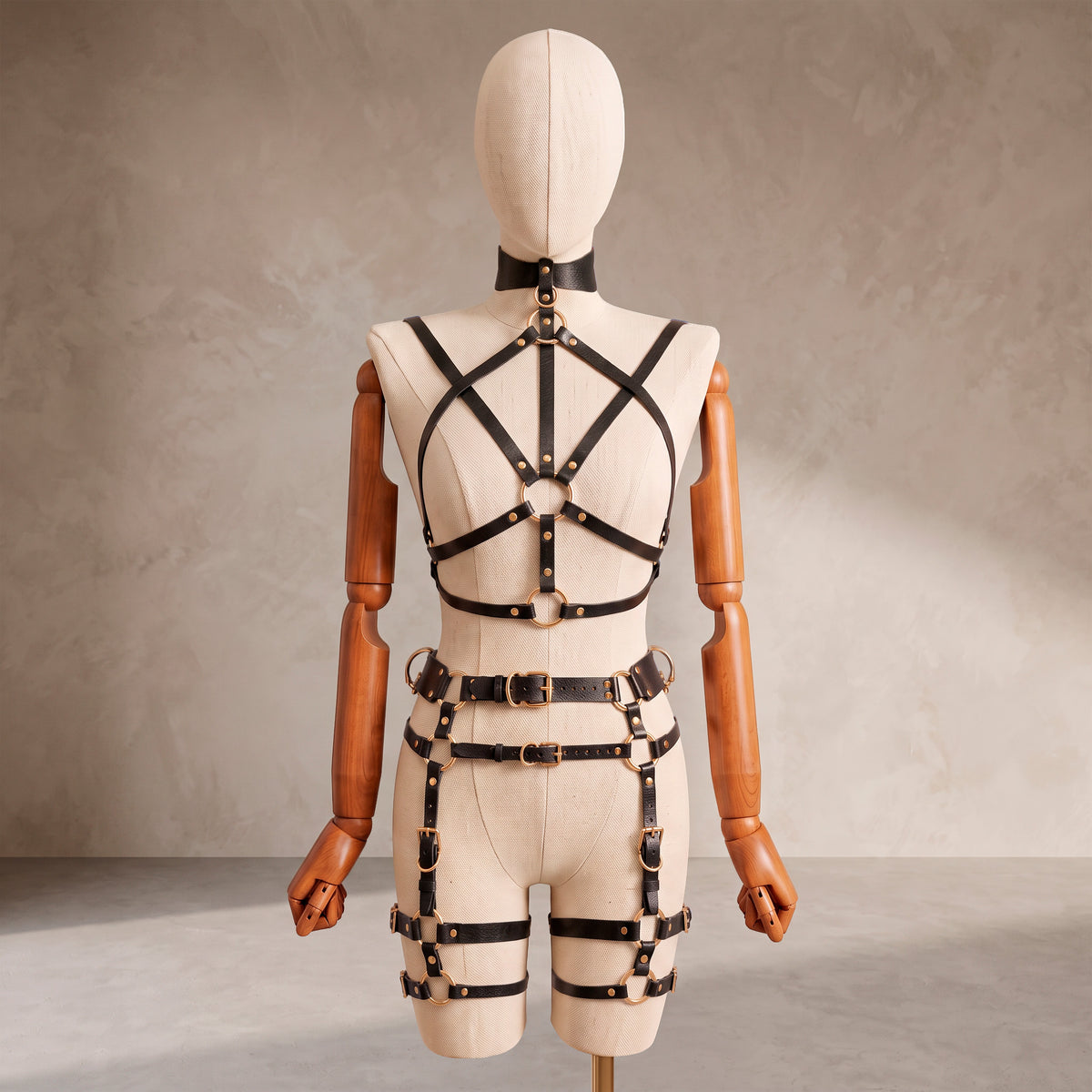 A cream-colored mannequin with wooden arms and head is shown from the back, wearing a black strappy harness-style bodysuit with gold-tone metal hardware against a textured beige studio background.