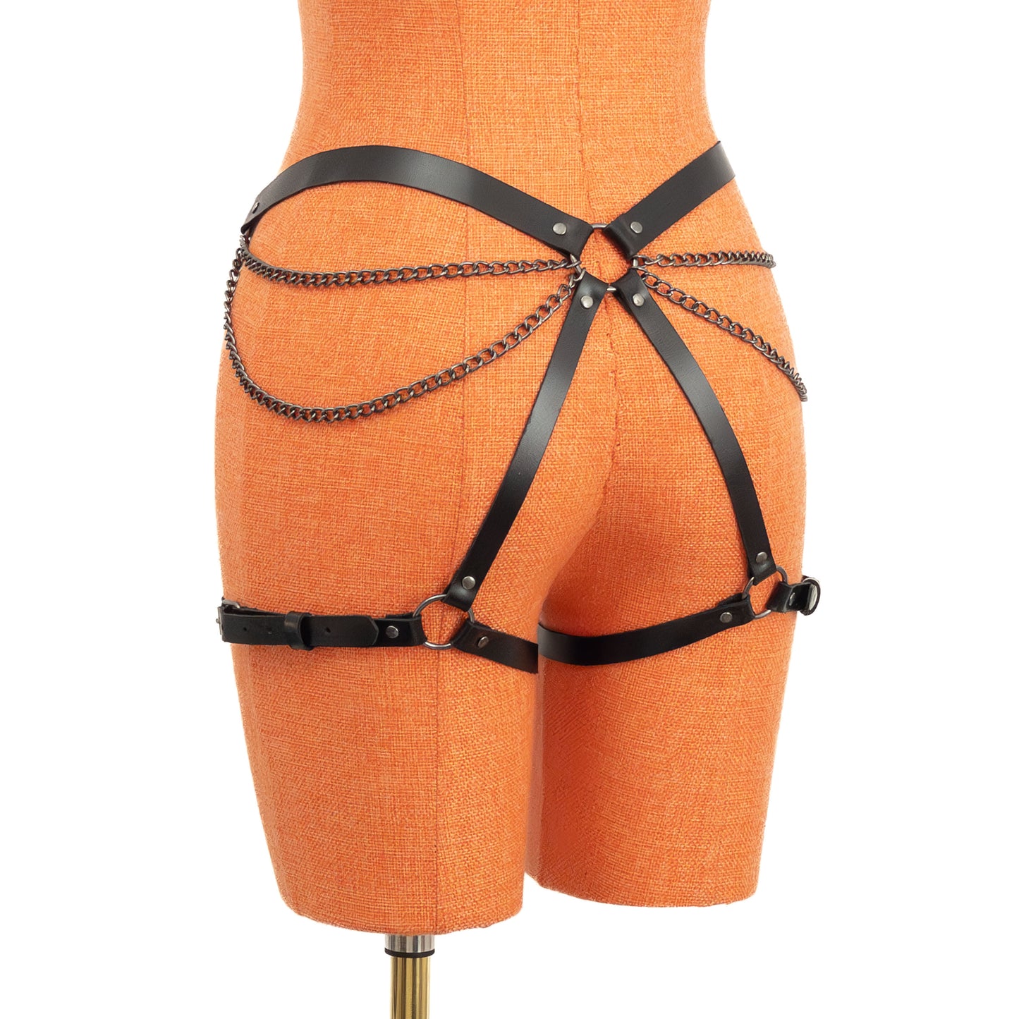 Angled rear view of the black genuine leather butt garter on an orange mannequin. The chain details create a layered pattern across the butt and hip line, meeting at the central back ring, while narrow thigh straps wrap evenly around both legs.