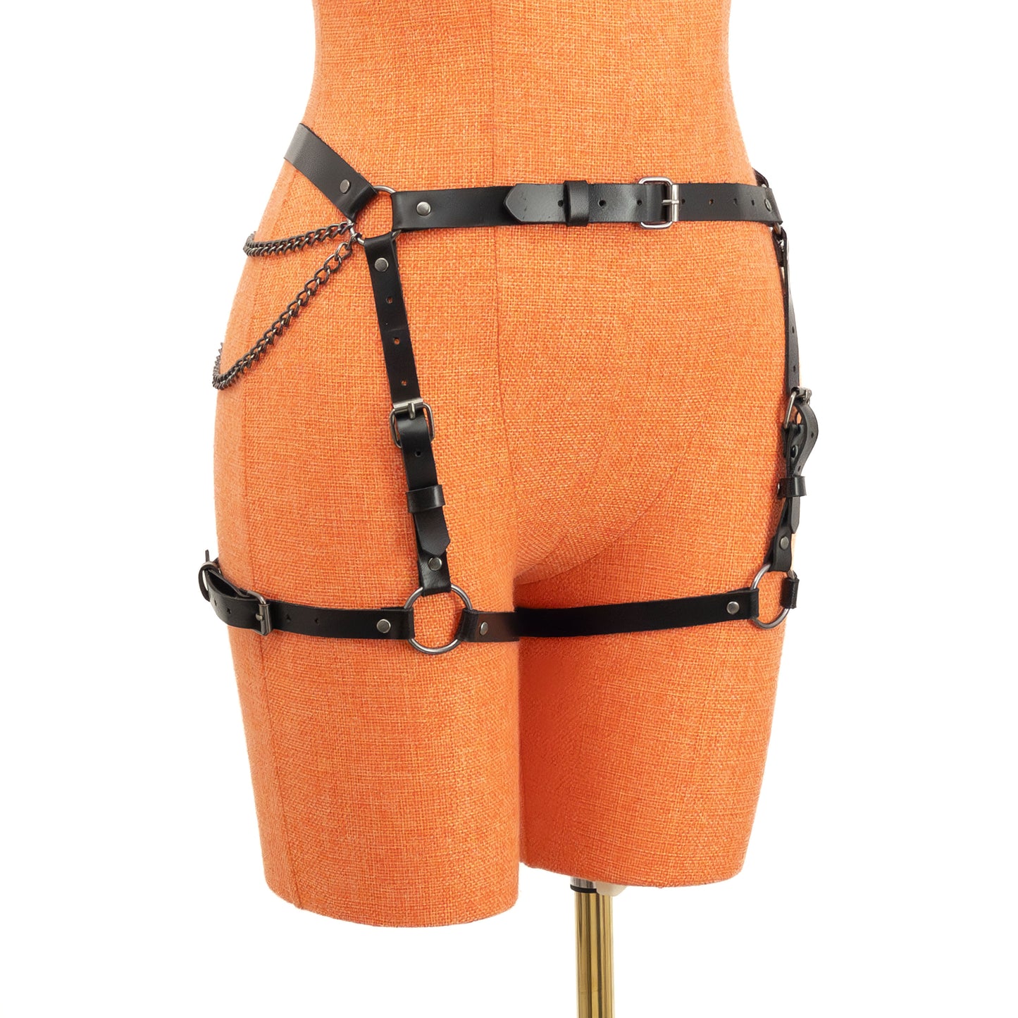 Semi-side view of the black leather butt garter on an orange mannequin. The narrow 5/8 inch (15 mm) waist strap, vertical front adjustment straps, horizontal thigh straps, and chain details leading to the central back ring are visible along the outer hip and thigh.