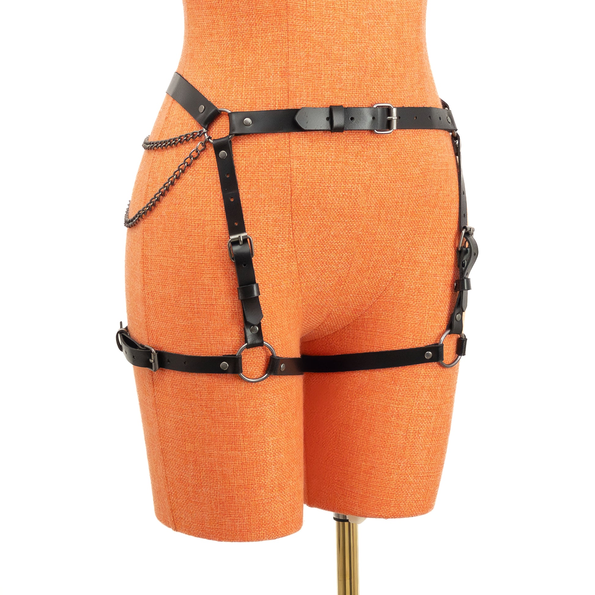 Semi-side view of the black leather butt garter on an orange mannequin. The narrow 5/8 inch (15 mm) waist strap, vertical front adjustment straps, horizontal thigh straps, and chain details leading to the central back ring are visible along the outer hip and thigh.