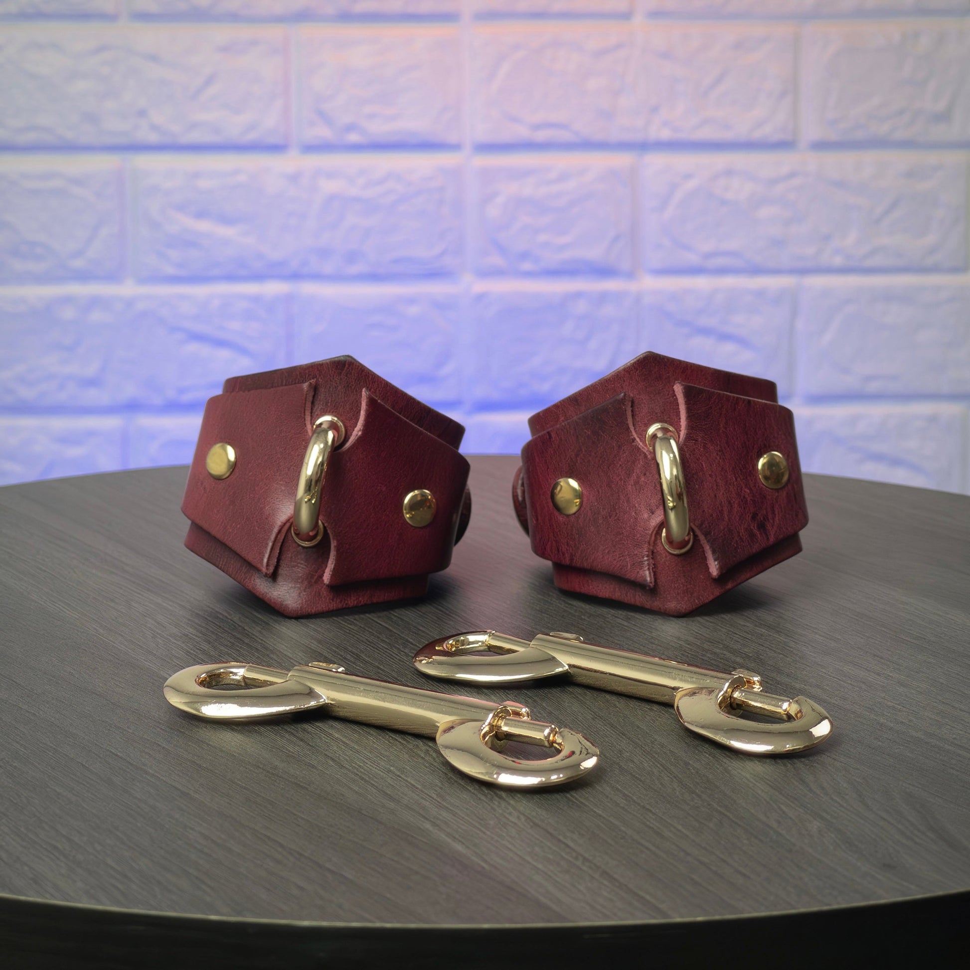 Two leather handcuffs with gold hardware on a dark surface against a textured light blue background.