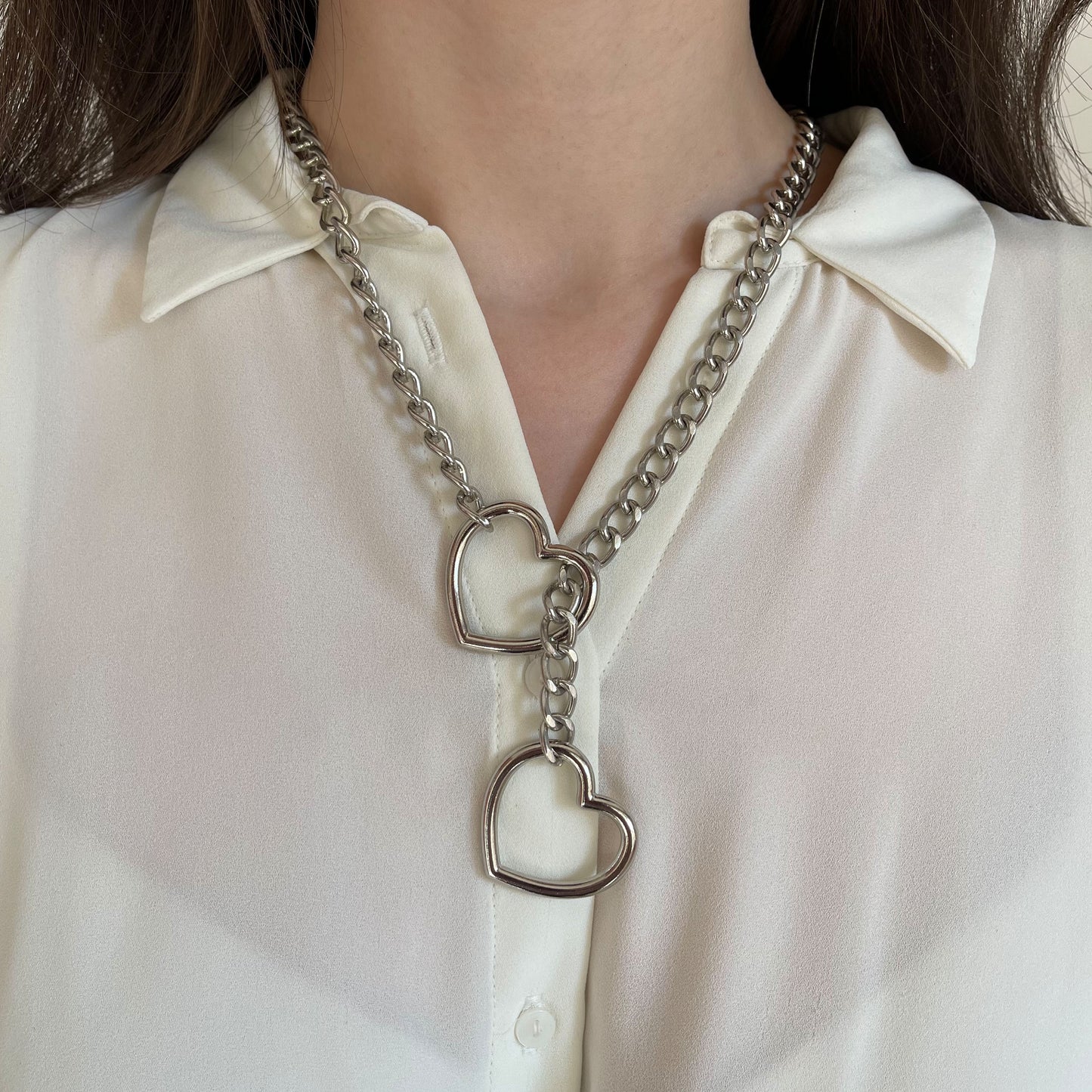 Silver sliding chain choking collar worn on the neck, heart-shaped ring at the front with the chain threaded through it, second heart ring hanging lower on the chest, metal chain forming a snug line around the neck, shown from the front.
