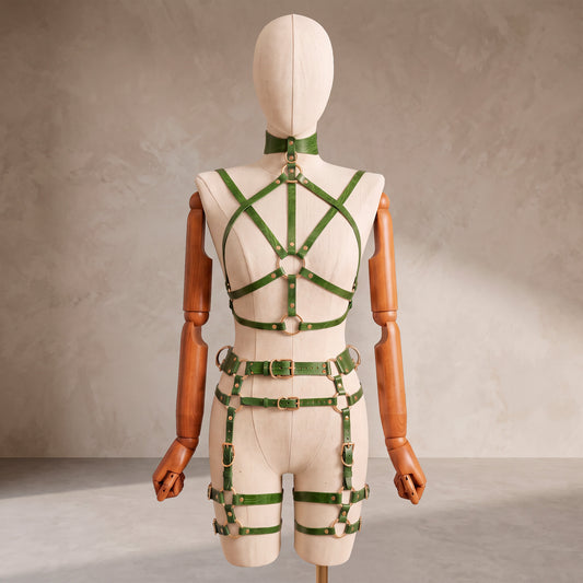 A cream-colored mannequin torso with wooden arms displays a green leather harness-style bodysuit with gold-tone metal rings and buckles, photographed against a textured beige studio backdrop.