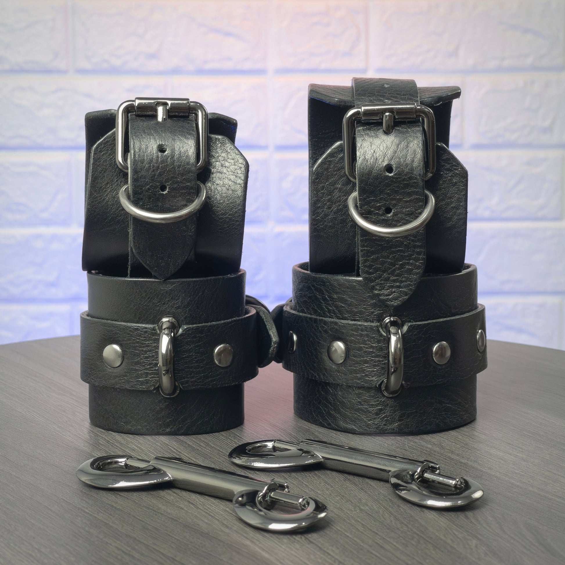 Black leather handcuff and ankle cuffs with silver hardware on a gray surface.