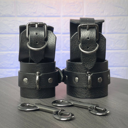 Black leather handcuff and ankle cuffs with silver hardware on a gray surface.