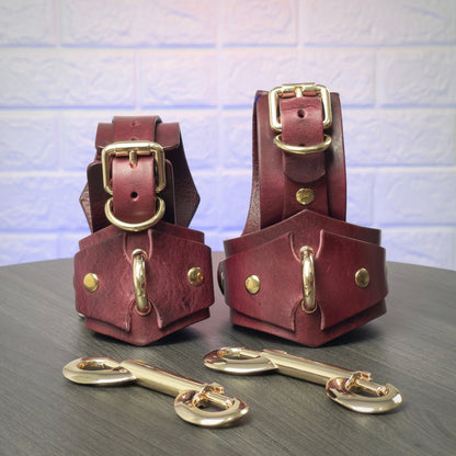 Pair of leather handcuffs and ankle cuffs with gold hardware on a wooden surface.