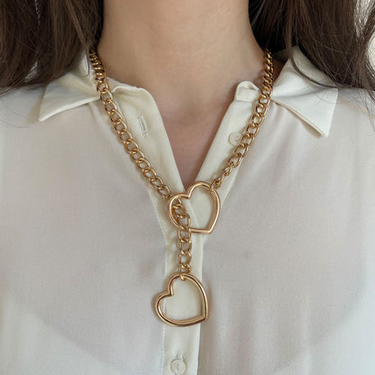 Gold sliding chain choking collar worn on the neck, heart-shaped ring positioned at the center of the chest, chain passing through the ring to create a tightening effect, second heart ring hanging below, smooth metal links visible.