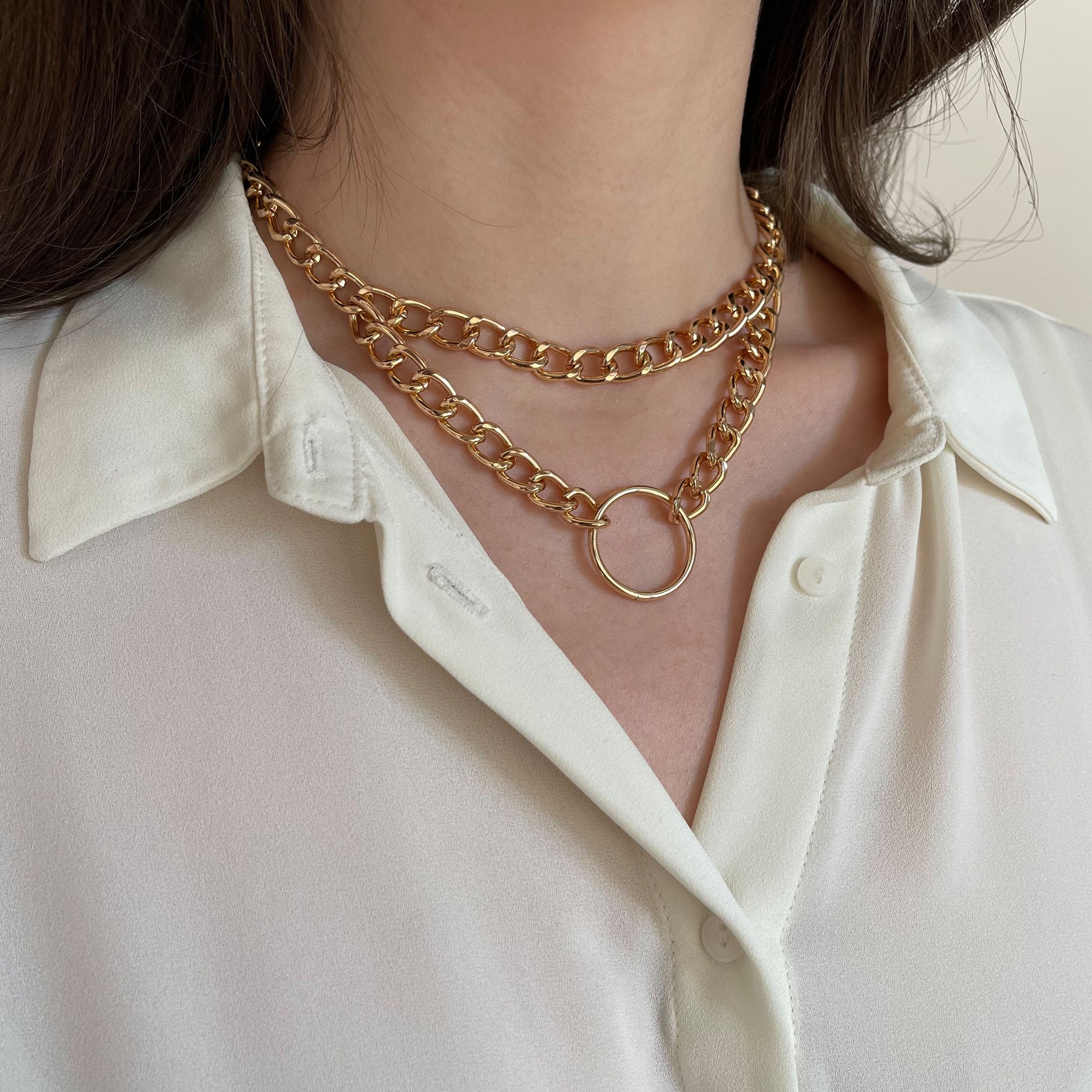 Double-layer gold-tone chain choker with a central metal ring. Worn on a woman’s neck over a white shirt, side angle shows the glossy finish and bold statement look. Lightweight and adjustable, this collar-style necklace blends minimalism with a touch of edgy elegance.