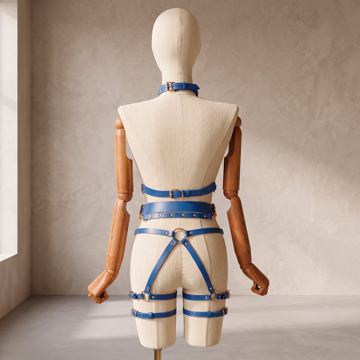Back view of a deep blue leather full body  harness set with an open bust harness, open crotch garter belt, removable leather handle, large front ring, adjustable straps, and gold-tone metal hardware.
